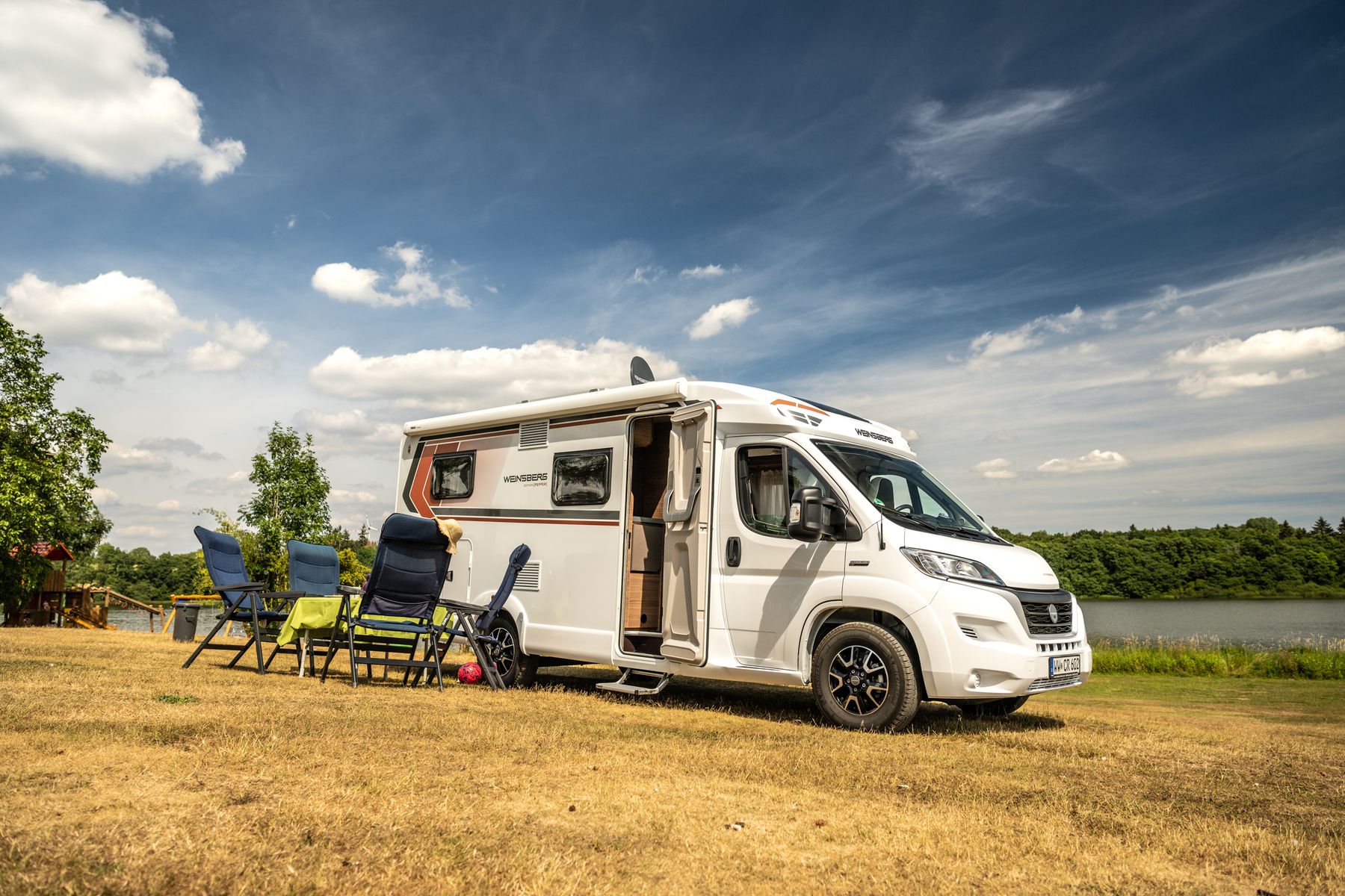 Ein weißes Wohnmobil ist auf trockenem Gras in der Nähe eines Sees unter einem teilweise bewölkten Himmel geparkt. Draußen sind Campingstühle und ein Tisch aufgestellt, im Hintergrund sind Bäume und Wasser zu sehen, was auf eine entspannende Szene im Freien hindeutet.